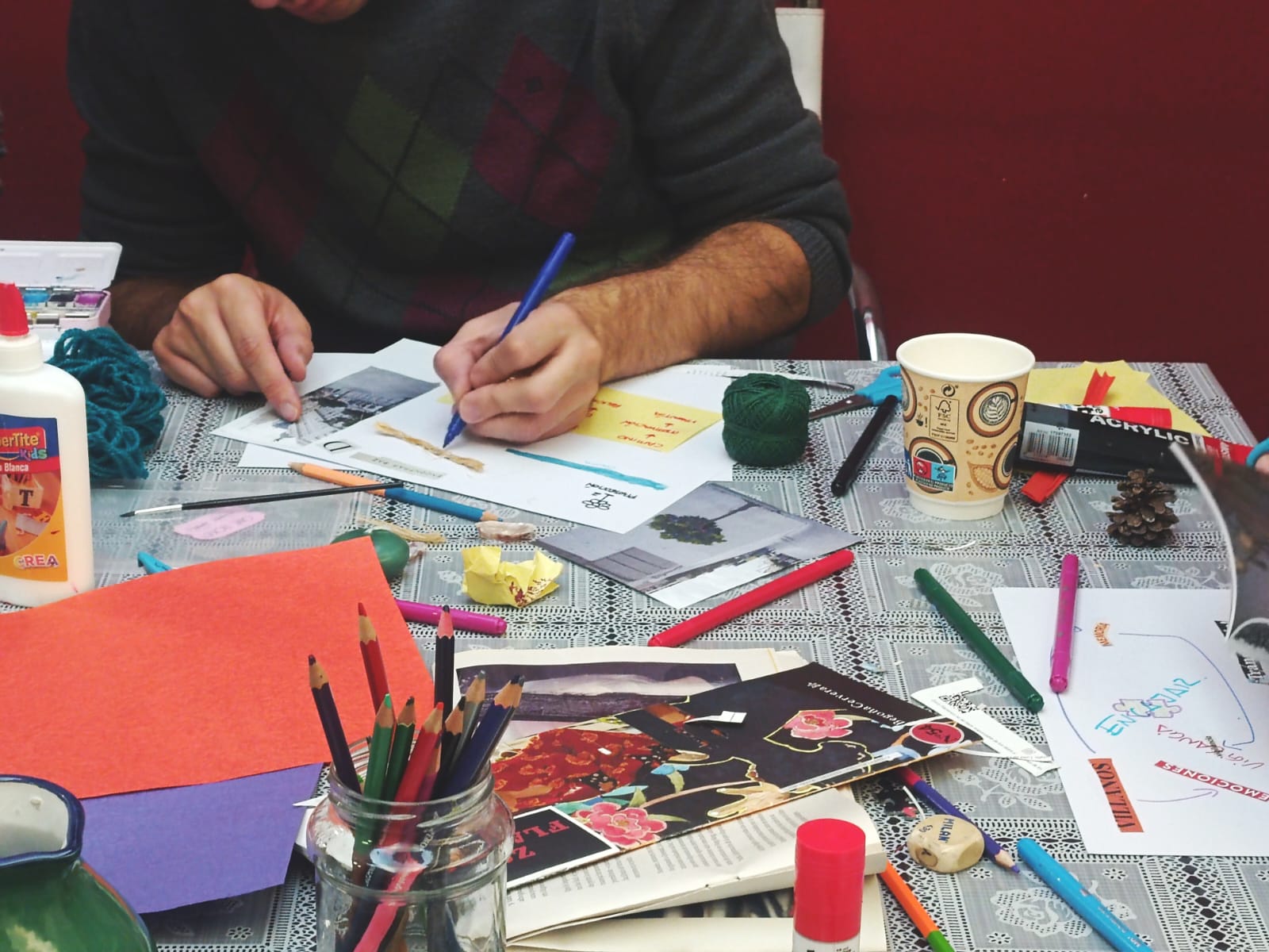 Persona dibujando sobre papel en una mesa llena de lápices, lanas y recortes